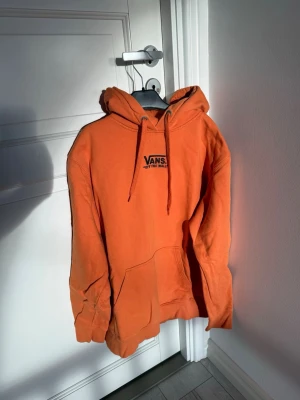 Orange hoodie från Vans - Snygg orange hoodie från Vans med svart logga och texten 'Off The Wall' på bröstet. Klassisk känguruficka framtill och justerbar huva med dragsko. Perfekt för en avslappnad streetwear-look.