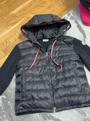 Svart pufferjacka från Moncler limited jacka - Snygg svart pufferjacka från Moncler med stickade ärmar och huva. Jackan har dragkedja framtill, två fickor och coola detaljer i rött, vitt och blått på snörena. Perfekt för kalla dagar och riktigt trendig look.