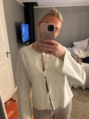 Vit kofta med knytband framtill - Trendig vit kofta med tre knytband framtill och långa ärmar. Koftan har en rak passform och är tillverkad i ett mjukt, tunt material som ger en avslappnad look. Perfekt att styla över ett linne eller topp.