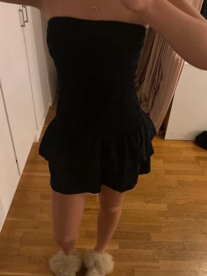 Svart bandeau jumpsuit med volang - Säljer en svart bandeau jumpsuit med smockad överdel och volangdetalj vid midjan. Kort modell med avslappnad passform och mjukt tyg som känns skönt mot huden. Perfekt för dig som vill ha en enkel men snygg look.