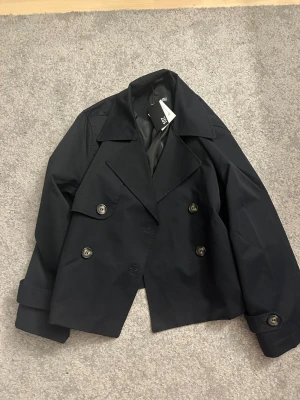 Svart kort trenchcoat från lager157 - Stilren svart trenchcoat från 157 med dubbelknäppning och bred krage. Jackan är kortare i modellen och har stora knappar samt klassiska detaljer. Tillverkad i ett slätt tyg med foder på insidan. Perfekt för dig som gillar minimalistisk och modern stil.