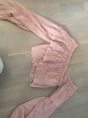 Rosa offshoulder volangblus - Supersöt rosa offshoulder-blus med volang och långa puffiga ärmar. Blusen har smockad överdel och är tillverkad i ett lätt, luftigt tyg som passar perfekt till sommaren. Ger en romantisk och trendig vibe till din outfit. Den har en flex som inte går bort men den syns inte 
