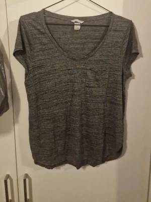 Grå melerad t-shirt med v-ringning - En enkel och snygg grå melerad t-shirt med v-ringad hals och korta ärmar. T-shirten har en liten bröstficka och är tillverkad i ett mjukt och tunt material som känns skönt mot huden. Perfekt till jeans eller shorts.