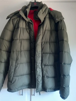 Olivgrön pufferjacka med huva - Säljer en olivgrön pufferjacka med avtagbar huva och röd fleecefoder upptill. Jackan har quiltad design, hög krage och tryckknappar framtill. Perfekt för kalla dagar och riktigt skön att bära. Materialet är syntet och jackan är vadderad för extra värme.