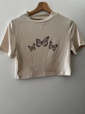 Beige croppad topp med fjärilar - Snygg croppad topp i beige med svart fjärilstryck framtill. Kortärmad modell med ribbad struktur och högre rund halsringning.