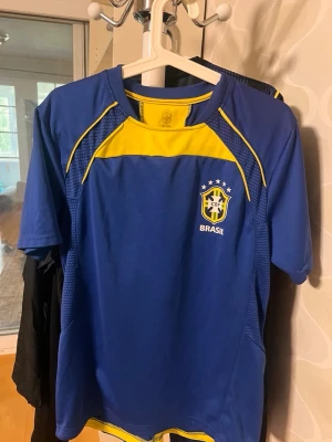 Blå Brasilien fotbollströja - Snygg blå fotbollströja med gula detaljer och Brasilien-emblem på bröstet. Tröjan har rund hals, korta ärmar och är tillverkad i ett lätt och ventilerande material som passar perfekt till träning eller match.