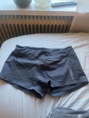 Mörkgrå träningsshorts från Virus - Snygga mörkgrå träningsshorts från Virus med bred midjeresår och logga tryckt på både framsidan och baksidan. Shortsen har en tight passform och är tillverkade i ett stretchigt funktionsmaterial som passar perfekt för gym eller löpning.