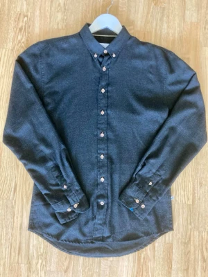 Kronstadt skjorta - Snygg svartgrå skjorta från Kronstadt i storlek L. Tillverkad i 65 % bomull och 35 % polyester, vilket gör den både bekväm och lättskött. Tyget har en mjuk, lätt borstad yta som ger ett stilrent intryck.  Skjortan är i fint begagnat skick utan hål eller fläckar. Endast små tecken på användning.  Detaljer:  * Märke: Kronstadt * Storlek: L * Färg: svartgrå * Material: 65 % bomull, 35 % polyester * Knäppning med knappar framtill * Button down-krage  Perfekt både till vardags och för en mer uppklädd