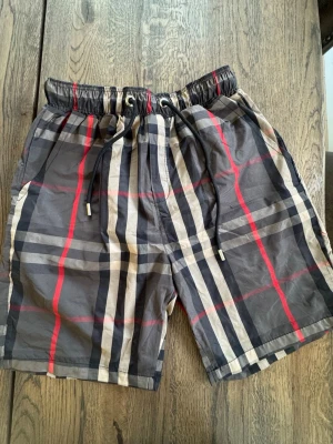 Rutiga shorts med snörning - Snygga shorts med klassiskt rutigt mönster i svart, grått, vitt och röda detaljer. Shortsen har elastisk midja med snörning och sidofickor. Perfekta för sommardagar och avslappnad stil.