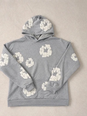 Grå hoodie med vita molntryck - Säljer en ljusgrå hoodie med huva och stora vita molnliknande tryck över hela tröjan. Hoodien har en klassisk känguruficka framtill och ribbade muddar vid ärmslut och nederkant. Perfekt för dig som gillar en unik och lekfull stil. Den sitter som M med så fungerar både för dem som har S och m 