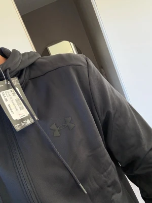Svart hoodie från Under Armour - Säljer en svart hoodie från Under Armour med dragkedja och justerbar huva. Hoodien har en diskret logga på bröstet och är tillverkad i mjukt fleece-material som känns riktigt skönt mot huden. Perfekt för dig som gillar sportig och clean stil. ALDRIG ANVÄND MED PRISLAPP KVAR