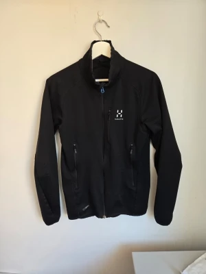 Snygg svart softshell kofta  - Snygg svart kofta från Haglöfs med hög krage och hel dragkedja. Jackan har två sidofickor med dragkedja och en bröstficka. Tillverkad i ett lätt och slitstarkt material.