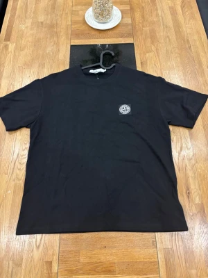 Stone Island  - Snygg svart t-shirt med rund hals och korta ärmar. På vänster bröst sitter en liten broderad logga som ger en clean och stilren look. Perfekt till jeans eller shorts för en enkel men trendig outfit.