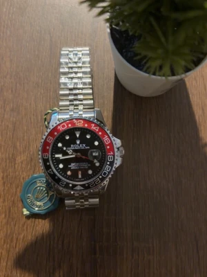 Klocka  - Klocka GMT-Master II med röd och svart vridring, svart urtavla och silverlänk i rostfritt stål. Klockan har tydliga index, datumfönster och klassisk jubilee-länk. Perfekt för dig som gillar exklusiva accessoarer.