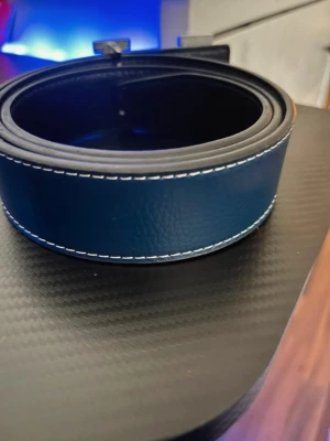 Herme’s bälte  - Snyggt blått bälte med vit kontrastsöm och stort H-format metallspänne. Bältet har en stilren design och är tillverkat i skinn, vilket ger en exklusiv känsla. Perfekt accessoar för att lyfta din outfit.
