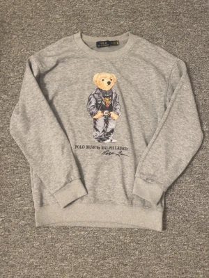 Grå sweatshirt Polo Bear Ralph Lauren - Säljer en grå sweatshirt från Ralph Lauren med den ikoniska Polo Bear-trycket på bröstet. Tröjan har rund hals, ribbade muddar och är gjord i mjukt material. Perfekt för dig som gillar streetwear och klassisk stil.