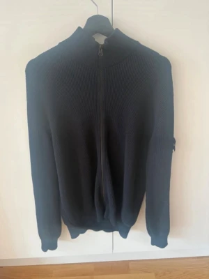 Svart ziptröja från Stone Island - Snygg svart stickad ziptröja från Stone Island med hög krage och ribbade muddar. Tröjan har dragkedja framtill och den klassiska Stone Island-loggan på ärmen. Perfekt för en stilren och avslappnad look.