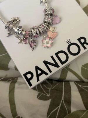 Pandora armband med rosa berlocker - Charmigt Pandora-armband i silver med flera berlocker i rosa och lila toner. Armbandet har hjärtan, blommor och gnistrande detaljer, vissa med emalj och pärlliknande inslag. Perfekt för dig som gillar personliga smycken med romantisk touch. Kvitton finns me