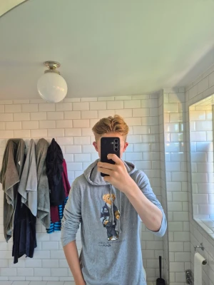 Grå hoodie Polo Bear från Ralph Lauren - Snygg grå hoodie från Polo Ralph Lauren med den ikoniska Polo Bear-trycket på bröstet. Hoodien har dragsko i huvan och är tillverkad i mjukt material som känns skönt mot huden. Perfekt för en avslappnad och trendig stil.