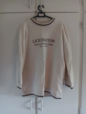Beige sweatshirt från Lexington - Snygg beige sweatshirt från Lexington med trycket 'American Heritage Est. 1993' på bröstet. Tröjan har bruna detaljer vid krage, ärmslut och nederkant. Perfekt för en avslappnad och stilren look. Använd fåtal gånger  är xs men passar även s/m 