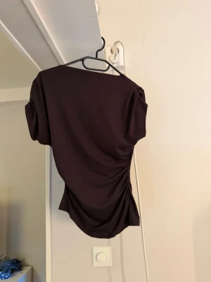 Mörkbrun topp  - Säljer en mörkbrun topp med kort ärm och snygg detalj på sidan. Toppen har en asymmetrisk form och är tillverkad i ett mjukt, stretchigt material som sitter bekvämt på kroppen. Perfekt för dig som vill ha en unik och trendig look.