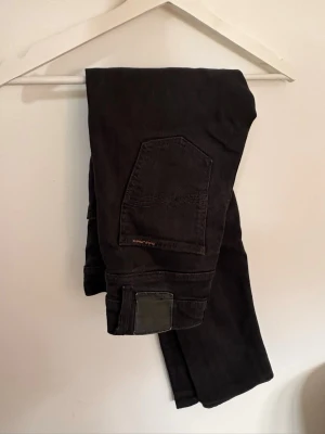 Svarta jeans från Nudie Jeans - Säljer ett par svarta jeans från Nudie Jeans i storlek 29/30. Jeansen har klassisk femficksdesign, raka ben och detaljerade sömmar på bakfickorna. Tillverkade i mjukt och slitstarkt denimtyg. Perfekta för en clean och stilren look.