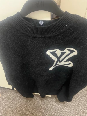 Svart stickad tröja med vit logga - Snygg svart stickad tröja med en stor vit logga på bröstet. Tröjan har rund halsringning och ribbade muddar vid halsen. Perfekt för dig som vill ha en enkel men trendig look till hösten eller vintern.