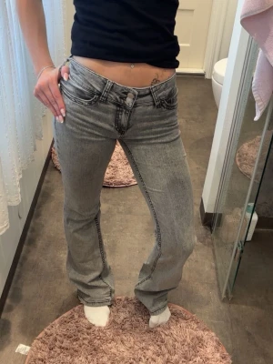 Grå bootcut jeans - Säljer ett par grå bootcut jeans med tydliga kontrastsömmar och låg midja. Jeansen har klassiska fem fickor och en snygg detalj på bakfickan. Perfekta för dig som gillar en avslappnad men trendig stil.