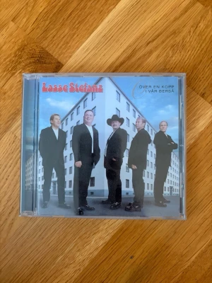 Lasse Stefanz CD original - Över en kopp i vår berså - CD-skiva med Lasse Stefanz, albumet 'Över en kopp i vår berså'. Omslaget visar bandmedlemmarna i svarta kostymer mot en bakgrund av ett höghus och blå himmel. Baksidan listar 15 låtar. Perfekt för dig som gillar dansband och svensk musik.