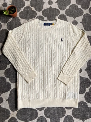 Vit kabelstickad tröja Polo Ralph Lauren - Kabelstickad vit tröja från Polo Ralph Lauren med klassisk marinblå logga broderad på bröstet. Tröjan har rund halsringning, långa ärmar och ribbade muddar. Perfekt för en clean och stilren look. XL fast känd som L