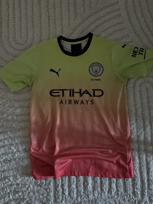 Manchester City matchtröja gul/rosa - Säljer en Manchester City matchtröja från Puma i gul och rosa gradient. Tröjan har svart krage, klubbmärke och sponsortryck på bröstet samt texten '125 YEARS'. Perfekt för dig som älskar fotboll och vill sticka ut på planen.