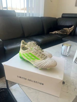 Vita och limegröna Adidas fotbollsskor - Säljer ett par riktigt snygga fotbollsskor från Adidas Limited Collection. Skorna är vita med limegröna detaljer och har klassiska tre ränder på sidan. De har snörning och en modern, strömlinjeformad design. Perfekta för dig som vill sticka ut på planen.