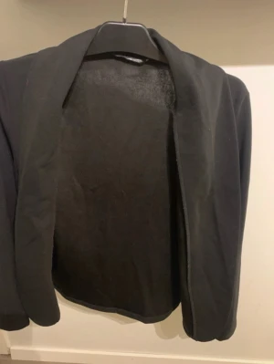Svart klassisk kavaj utan knappar - En stilren svart kavaj med öppen front och mjuk passform. Kavajen har en enkel design utan knappar och är perfekt för en clean och modern look. Materialet ser ut att vara ett mjukt tyg som ger en avslappnad känsla.