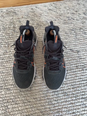 Svarta Nike sneakers med orange detaljer - Säljer ett par svarta Nike sneakers med orange swoosh och detaljer. Skorna har en sportig design med ventilerande meshmaterial och en vit sula med svarta inslag. Perfekta för dig som gillar streetstyle och vill sticka ut med snygga färgkontraster.