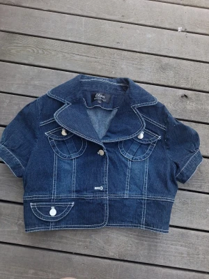 Kort jeansjacka med rosett bak - Snygg mörkblå jeansjacka från Elam med korta ärmar och markerade vita sömmar. Jackan har två bröstfickor med lock och dekorativa knappar samt en söt rosett i ryggen. Klassisk krage och kort, boxig passform som ger en trendig look.