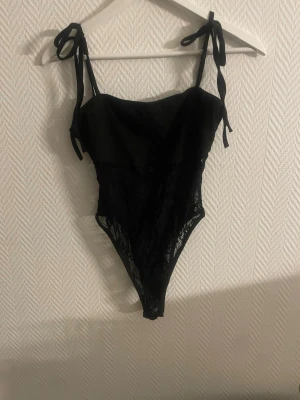 Svart spetsbody med knytband - Snygg svart body med tunna axelband som knyts på axlarna. Överdelen är i slät tyg medan nedre delen är i genombruten spets som ger en elegant look. Hög skärning vid benen och figurnära passform. Perfekt för dig som vill ha något stilrent och trendigt.