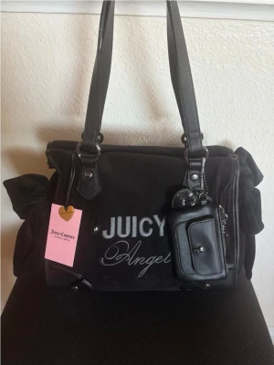 Juicy Couture svart velourväska - Juicy Couture handväska i svart velour med broderad text 'JUICY Angel' på framsidan. Väskan har dubbla axelband i svart konstläder, dekorativa rosetter på sidorna och en avtagbar liten accessoarväska framtill. Perfekt för dig som gillar Y2K-stil.