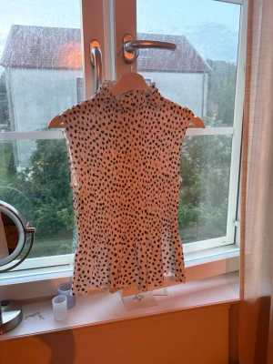 Prickig ärmlös topp med volang - Snygg vit ärmlös topp med svarta prickar och hög krage. Toppen har en lätt volang nertill och är tillverkad i ett tunt, luftigt material som passar perfekt till varma dagar. Den har en figurnära passform och ger en trendig look. Perfekt nu till sommaren andvänt få antal gånger 🥰 från NAKD   Pris kan förhandlas 