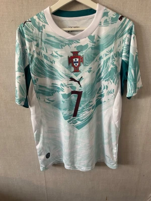 Portugal Ronaldo matchtröja Puma - Snygg Portugal matchtröja från Puma med Ronaldo och nummer 7 på ryggen. Tröjan är vit med turkosa mönster och har korta ärmar. Perfekt för dig som älskar fotboll och vill stötta Portugal på match eller träning.