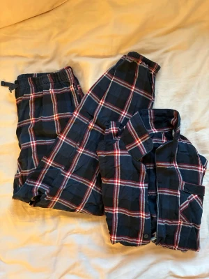 Mörkblå rutiga pyjamas i bomull - Klassisk tvådelad pyjamas med skjorta och byxa i mörkblått med rött och vitt rutmönster. Skjortan har krage och knappstängning, byxorna har resår i midjan. Tillverkad i mjuk bomull, perfekt för mysiga kvällar.