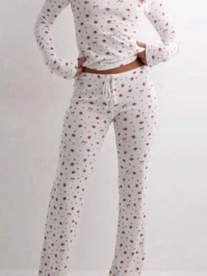Vit blommig med rosetter pyjamas - Supermysig vit pyjamas med små blommor och rosetter  i rött. Setet har långärmad tröja och byxa med snören i midjan. Perfekt för chillkvällar hemma. Mjukt material och relaxed fit.