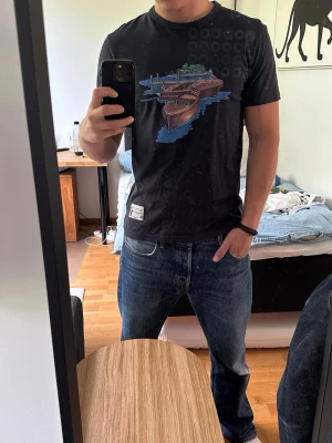 Svart t-shirt från Woolrich med tryck - Svart t-shirt från Woolrich i bomull med coolt tryck av en båt och naturmotiv på bröstet. Klassisk rund hals och korta ärmar. Perfekt för dig som gillar stilrena plagg med en twist.
