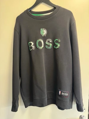Svart BOSS Celtics sweatshirt - Cool svart sweatshirt från BOSS med Boston Celtics-logga och gröna detaljer på bröstet. Tröjan har rund hals, långa ärmar och NBA-märke nedtill. Perfekt för dig som gillar basket och streetwear.