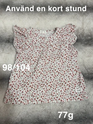 Blommig topp med volangärmar - Söt blommig topp i vitt med små röda och gröna blommor. Blusen har volangdetaljer vid ärmarna och rund halsringning. Perfekt för sommarens alla tillfällen.🌸 Använd en kort stund. Storlek 98/104