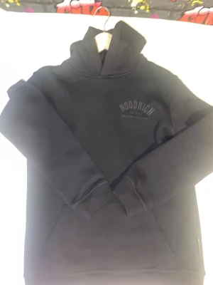 Svart hoodie från Hoodrich - Säljer en svart hoodie från Hoodrich med trycket 'HOODRICH EST. 2014 WELCOME TO THE FAMILY' på bröstet. Hoodien har huva och magficka, samt ribbade muddar vid ärmslut och nederkant. Perfekt för en avslappnad streetwear-look.