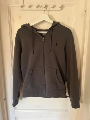 Mörkgrå hoodie från Polo Ralph Lauren - Säljer en snygg mörkgrå hoodie från Polo Ralph Lauren med dragkedja, huva med dragsko och klassisk logga broderad på bröstet. Mjuk bomullsblandning och två stora fickor framtill. Perfekt för en avslappnad och stilren look. Hör av dig vid frågor🙌🏼