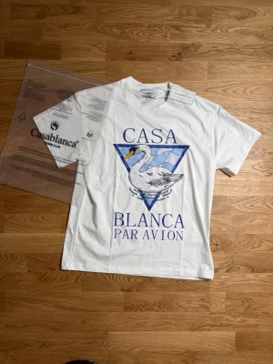 Vit Casablanca t-shirt med svanmotiv - Casablanca t-shirt i vitt med stort tryck framtill som visar en svan och blå triangulärt mönster. Texten 'Casa Blanca Par Avion' i blått. Klassisk passform och rund hals. Perfekt för dig som gillar stilrena men ändå unika plagg.