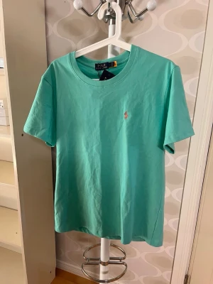 Turkos t-shirt från Polo Ralph Lauren - Snygg turkos t-shirt från Polo Ralph Lauren med klassisk passform och liten broderad logga på bröstet. Tillverkad i mjuk bomull och har rund halsringning samt korta ärmar. Perfekt för en avslappnad och stilren look.