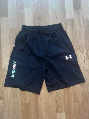 Under Armour träningsset svart/vit - Snyggt träningsset från Under Armour med svarta shorts och vit t-shirt. Shortsen har elastisk midja och logga på benet, t-shirten har tryck på bröstet och sidan. Båda i lätt och snabbtorkande material, perfekt för träning.