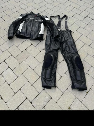 Svart och vit mc-skinnställ Dainese - Säljer ett svart och vitt mc-skinnställ från Dainese i storlek 54. Setet består av jacka och byxor i äkta läder med skydd vid axlar, armbågar och knän samt avtagbara hängslen. Perfekt för dig som vill ha stil och säkerhet på hojen.